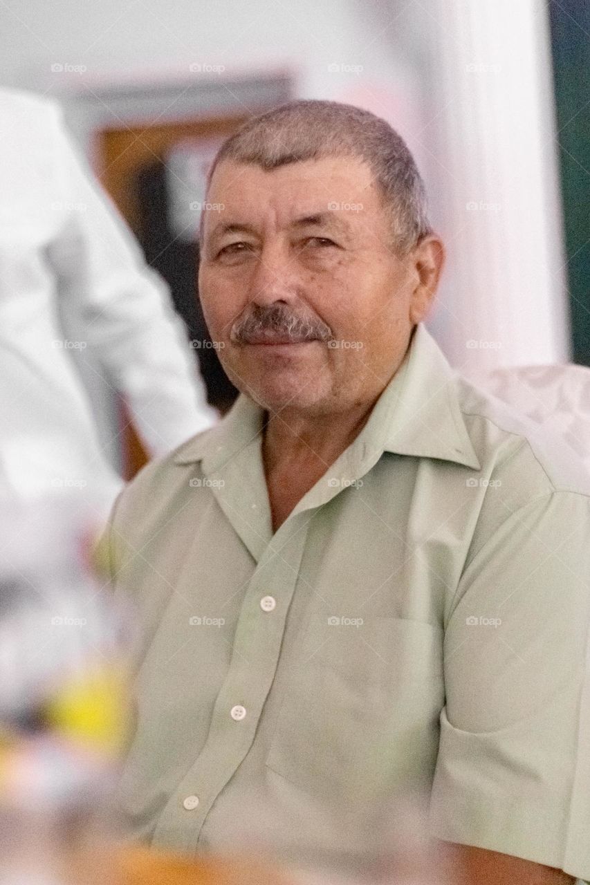 Portrait of an elderly Caucasian male brunet with a mustache looking sweetly at the camera with a smile, sitting on a chair in a room during the day, close-up view from the side.