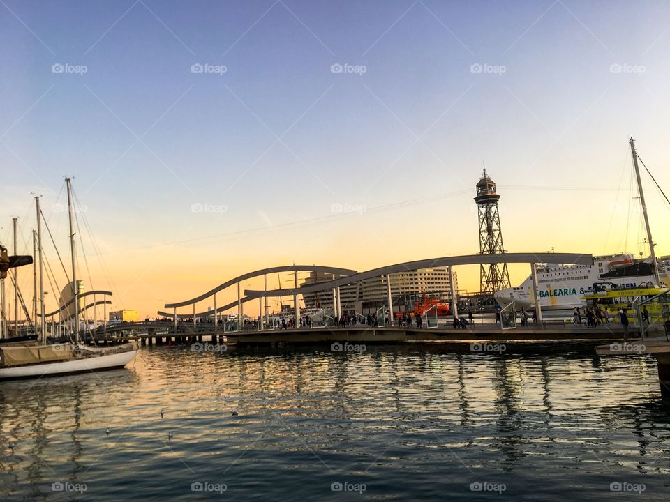 Golden time harbor, there are many people walking on a very beautiful bridge. There are some sailing boats and yachts moored , golden light shone into the cool water that look so wonderful. The sky is light blue and cloud below is pale yellow