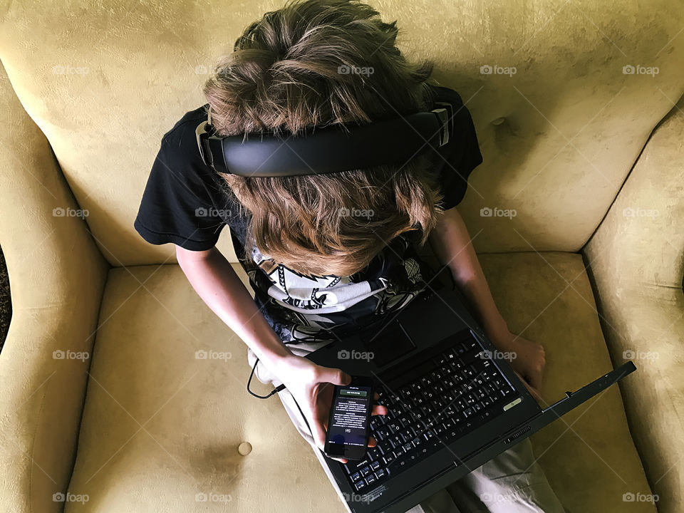 Overhead view of a teenager with headphones using a laptop, mobile phone and listening to the music on the yellow sofa at home 