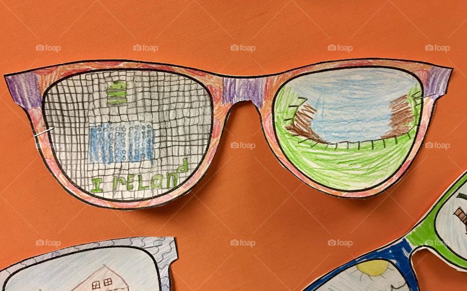 Several paper sunglasses, hand-drawn by a child, with colorful pictures on the lenses, placed against a bright orange construction paper background. The playful designs reflect a fun and creative artistic touch.