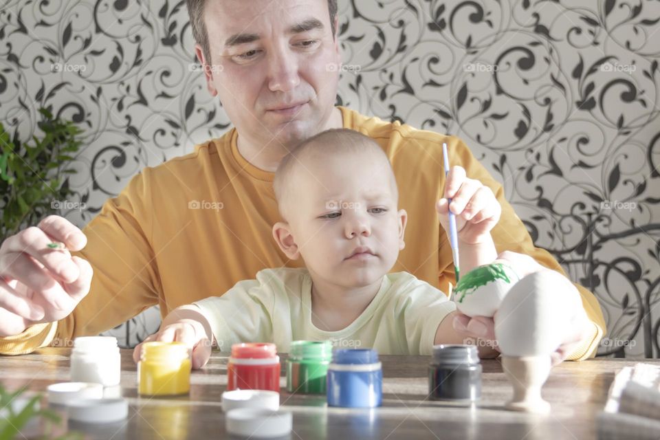 A little son and his father together in their free time paint Easter, chicken eggs with paints and brushes for the holiday. A child and a man create a craft and natural product in their family business.