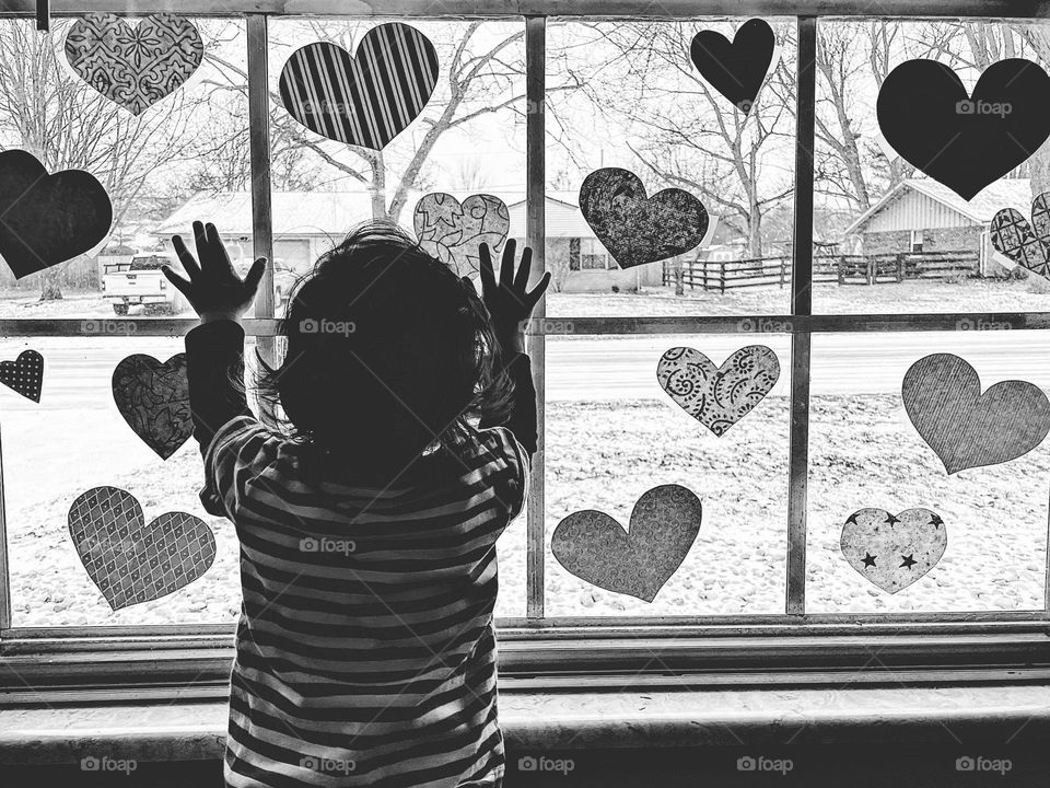Baby at window with hands reaching out for heart decor, baby looks out window with Valentine’s Day decorations on it, watching the snow, baby looks out of window, black and white portrait, monochrome image of baby in window, people in windows
