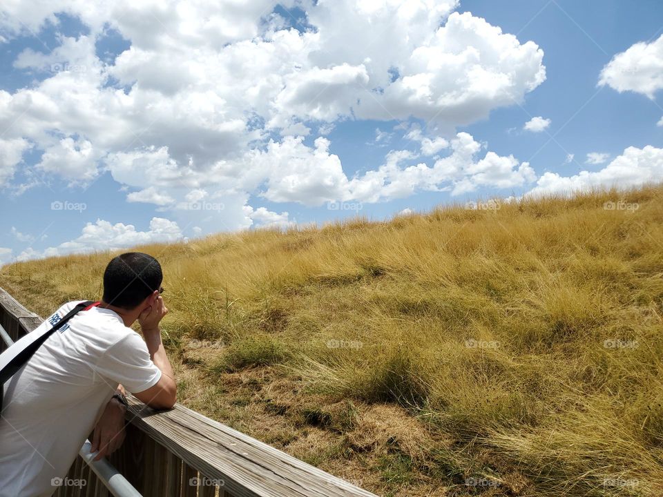 A man leaning on the rails of a wood deck path as he peacefully gazes out to the clouds beyond the golden grassy hill. He is alone and in a moment of stillness that transcends to meditative state that nature often brings.