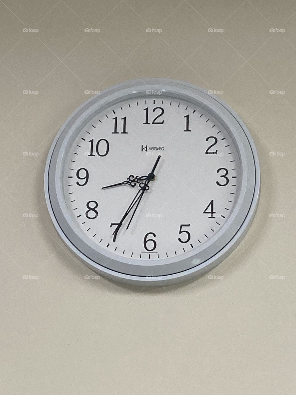 Relógio com bordas cinza , grande de parede, sobre uma parede cor de areia.