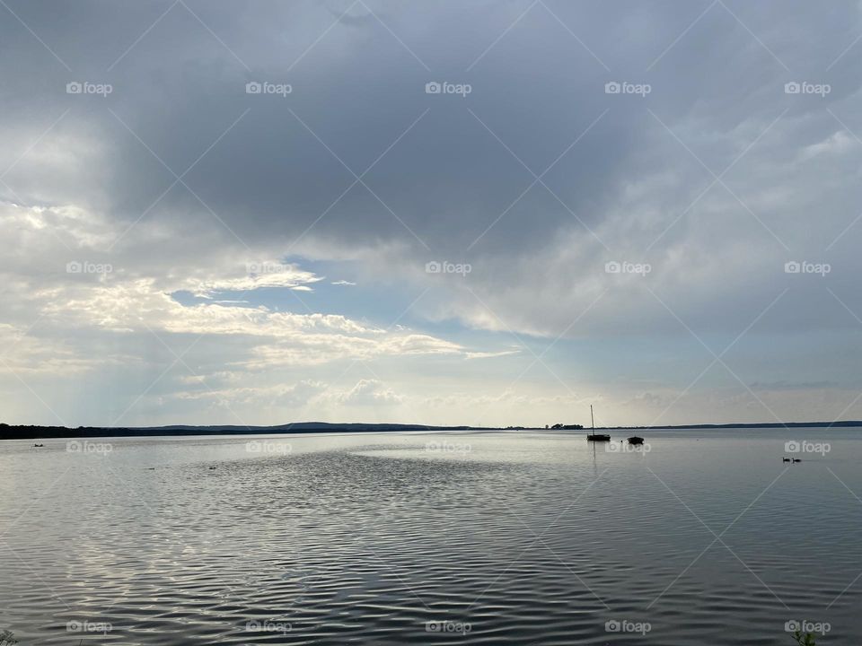 Aufziehender Sturm über dem See am Abend. Es liegen zwei Jollen auf dem See, die untergehende Sonne erahnt man noch am Horizont/ 