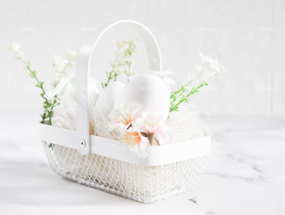 One white wicker metal basket with white straw, eggs and spring flowers stand on a white moamer table, close-up side view. Easter concept, white on white.