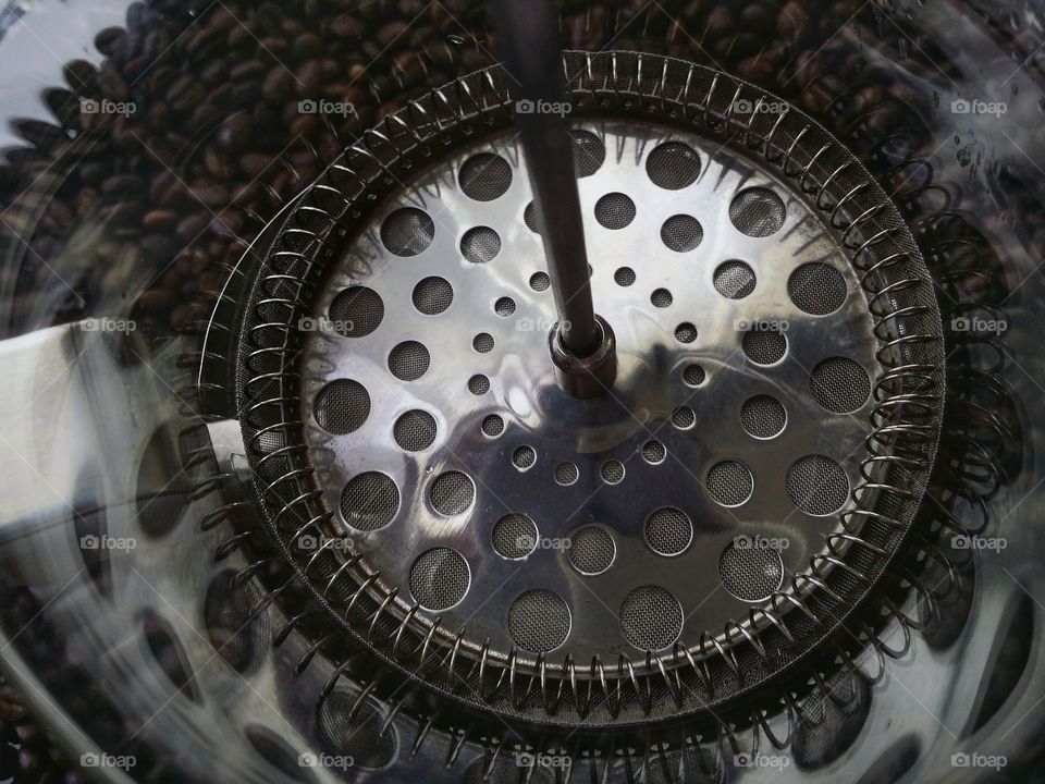 A french press coffee carafe from inside looking from above at the round circle of the strainer with whole coffee beans in the backgound