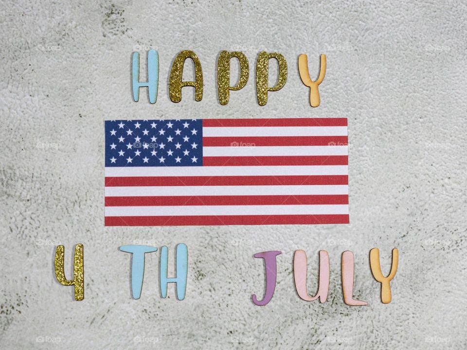 One paper American flag with colored shiny wooden letters with the words Happy 4th of July lying on a light stone background,flat Lay close-up.
