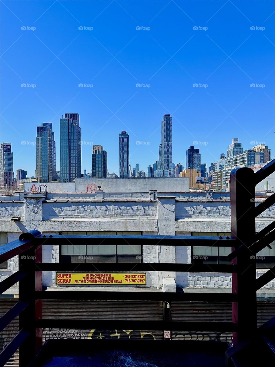 This is the red metal staircase of the “Pulaski Bridge” at “Newtown Creek” in LIC, Queens about half way up. The cityscape in the far background is “Manhattan”. 2024. Hypnotic Productions