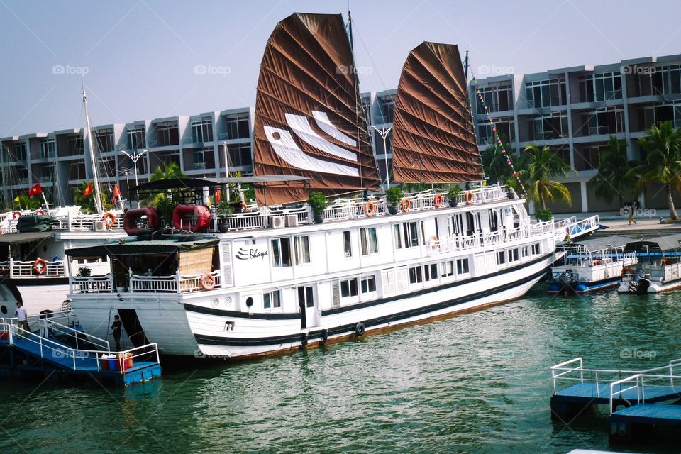 A white yacht with brown sails is moored on the dock, with windows, glass doors, curtains and decorated with potted plants, waiting for tourists