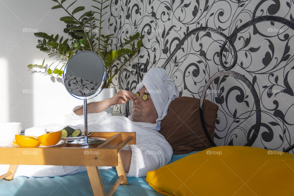 A man and home spa care at home takes care of himself and smears his face with cream, does a manicure at home on a blue background and with oranges and rests.