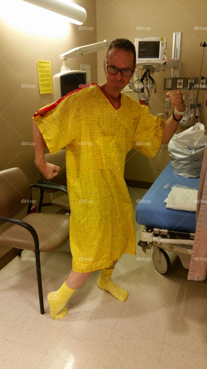 Man in hospital wearing a yellow hospital gown, flexing his muscles like he's invincible.