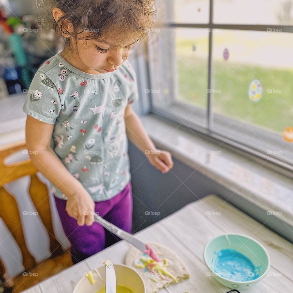 Toddler girl frosts Easter egg cookie, toddler decorating Easter egg sugar cookie, making sugar cookies for the holiday with toddlers, family traditions for Easter, fun with toddlers in the spring, in the kitchen with toddlers