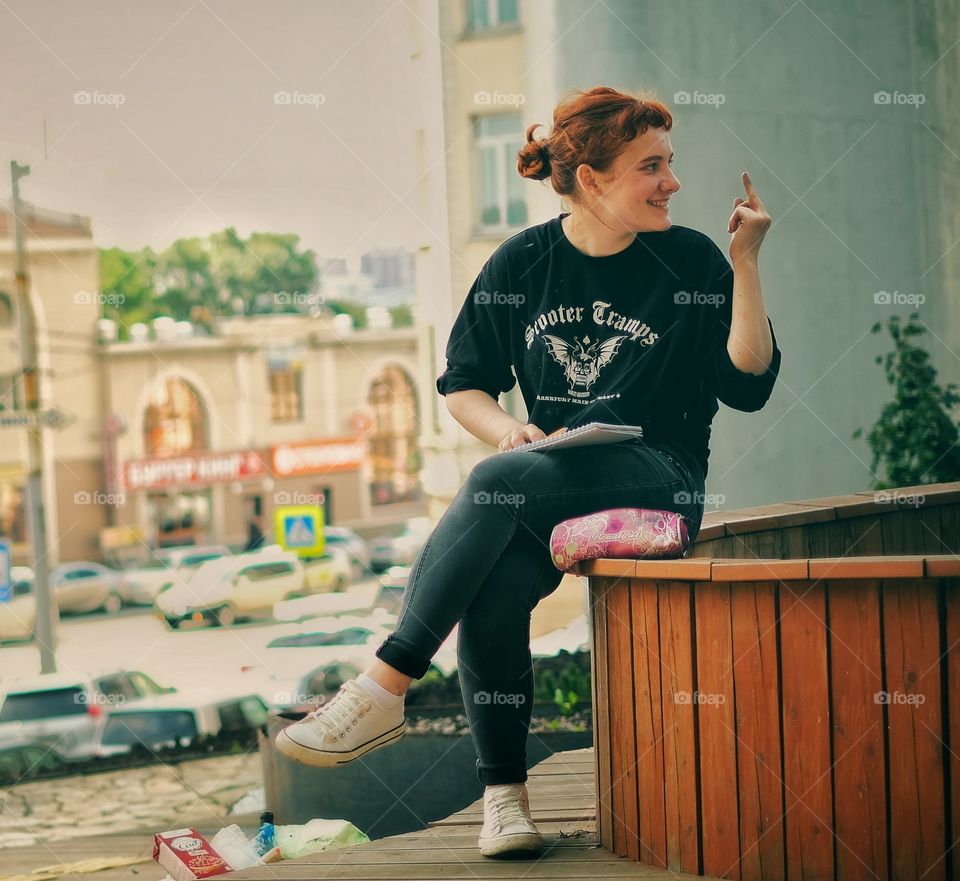 an artist girl wearing a black shirt, trousers, and white shoes who waa sitting in a park is stretching her middle finger by smiling.