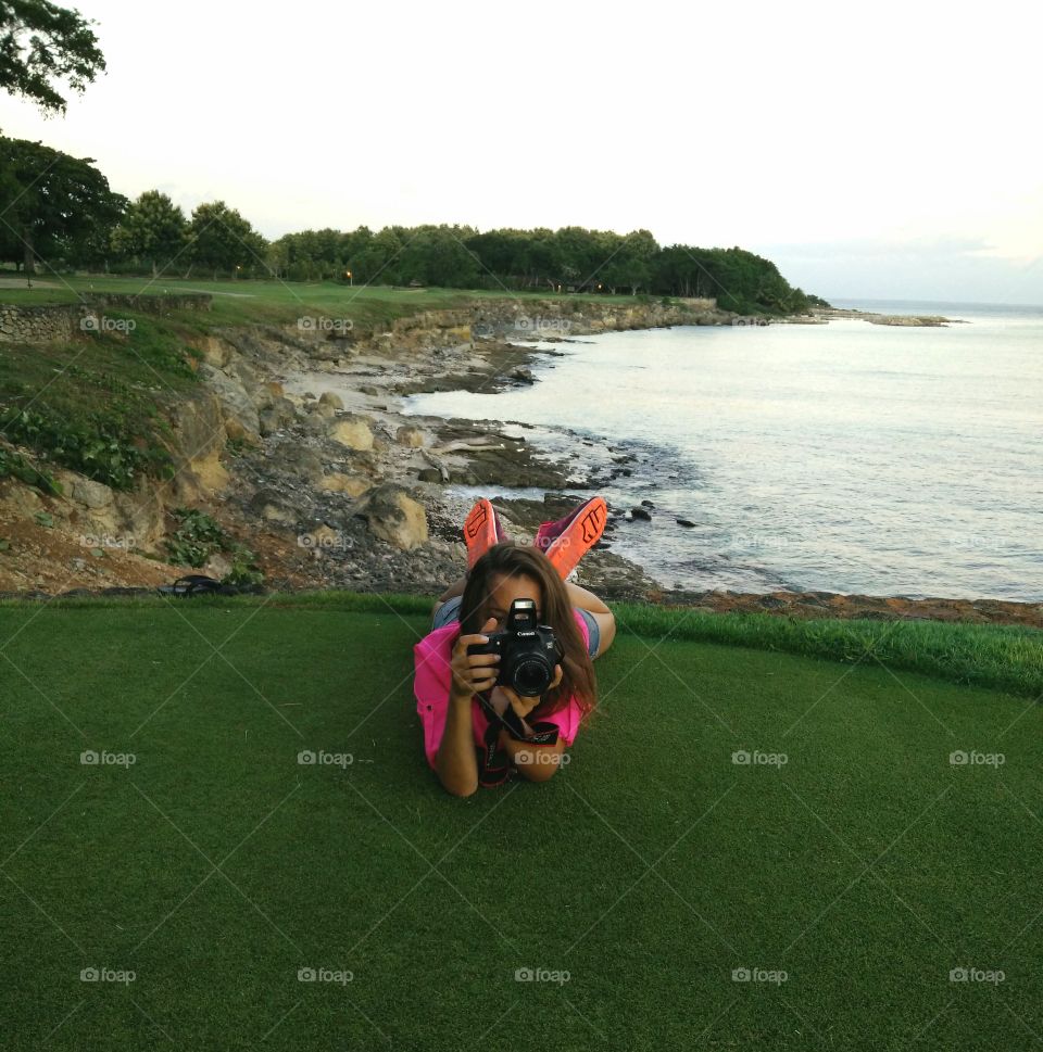 Young girl with a photo camera, making shots. Laying down on the golf field. Green color, and Caribbean sea.