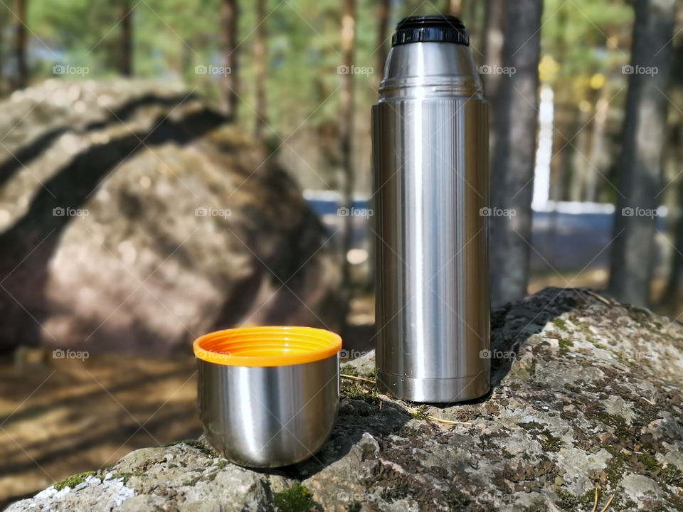 A thermos with hot tea or coffee stands on a large stone in the forest during a picnic or relaxing on a journey in the summer season.