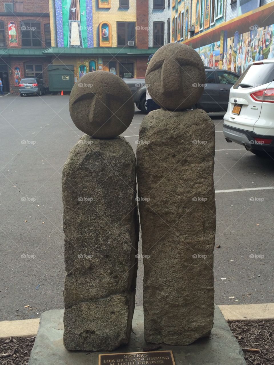 Stone sculpture of two figures in urban setting surrounded by parking lot, cars and murals