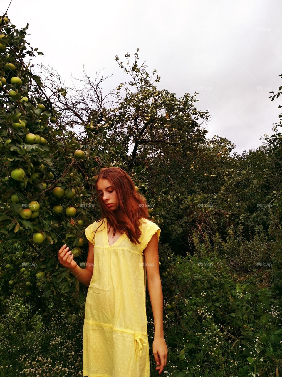 It is a photo of a young woman in the garden (apple trees). She has red (ginger) hair. She has brown eyes. She has medium lendth hair. She wears yellow dress