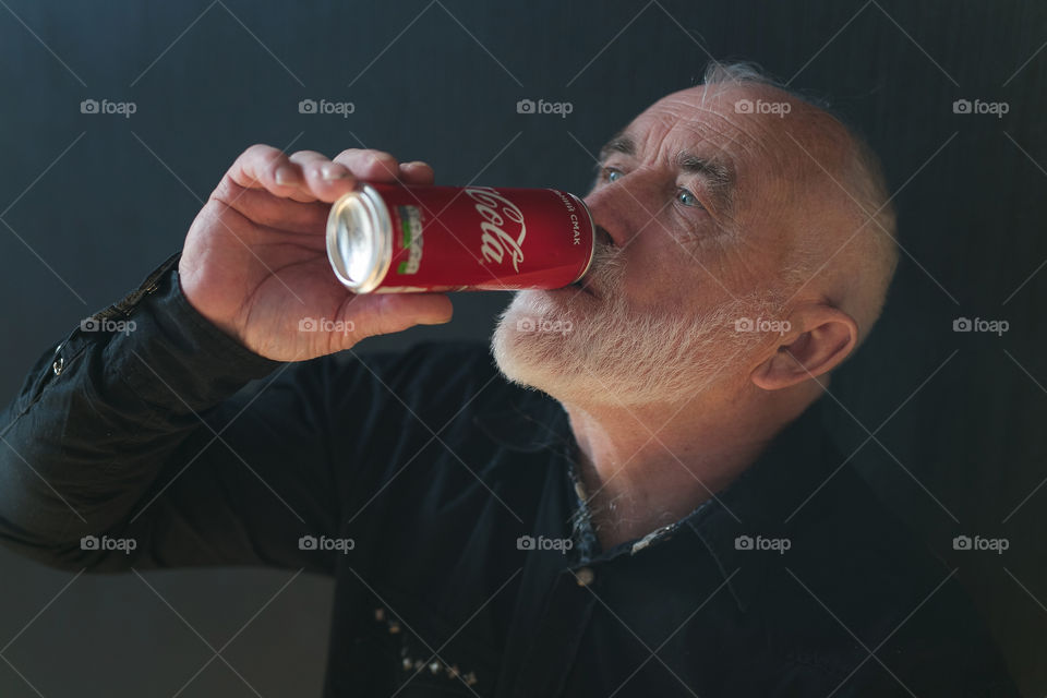 a handsome man with a gray beard and gray hair, in a black shirt, drinking Coca-Cola.  stylish man of about sixty