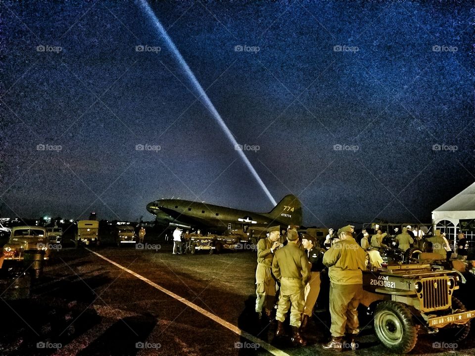 WWII Reenactment at night, bommers, airplanes, warbirds, military vehicles, people and soldiers with a bright searchlight in the black sky