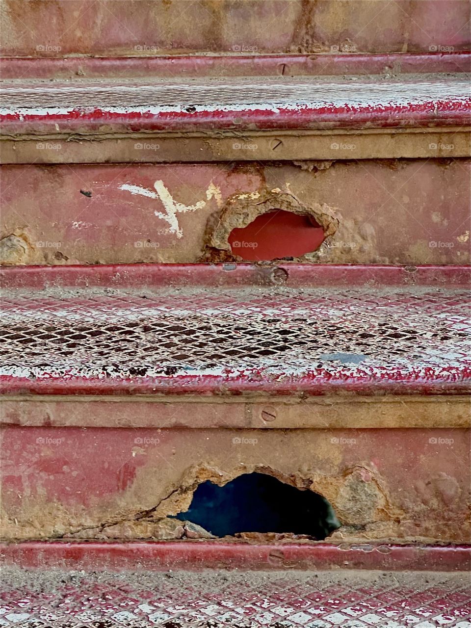 These are corroded holes in the red metal staircase of the “Pulaski Bridge” at “Newtown Creek” in “Greenpoint”, Bklyn. Thousands of people walk up these steps so the otherwise bright red has worn away to a tan maroon. 2024. Hypnotic Productions