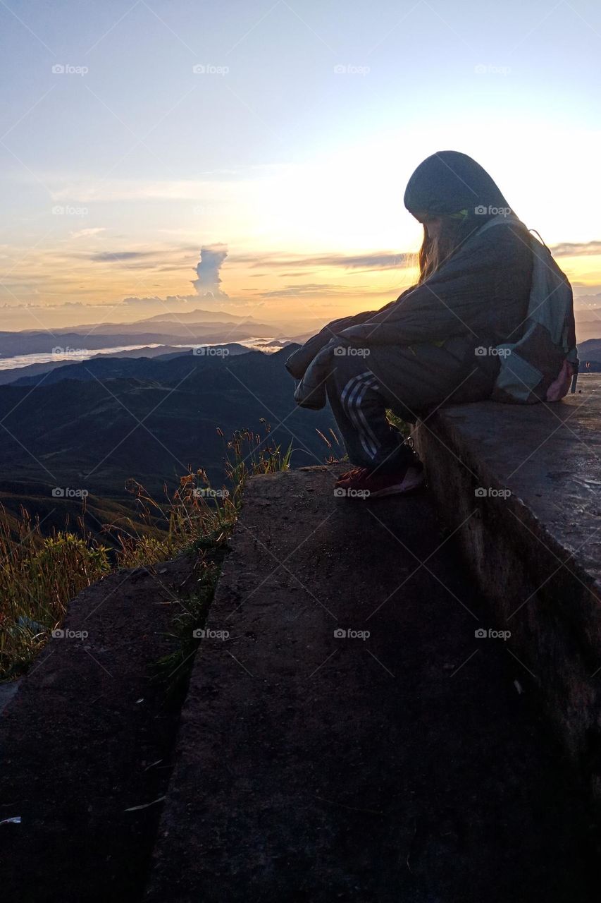 Sad girl sit on steps at the top of a high mountain during sunrise. She is feeling cold and carries a backpack.