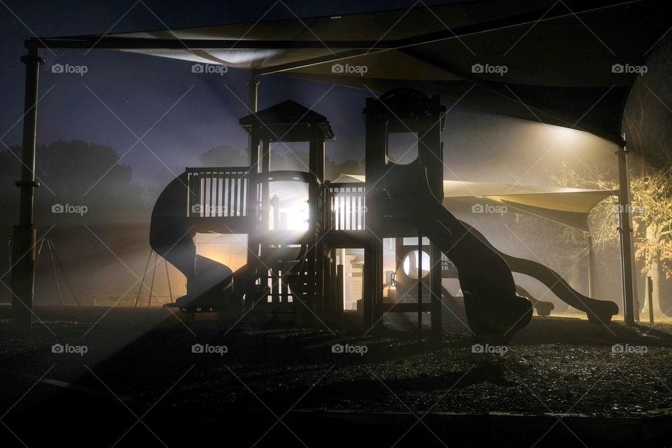 A surreal playground scene on a dark foggy morning, with the play equipment backlit by beams of light from the street lamps. Lake Benson Park in Garner North Carolina. 