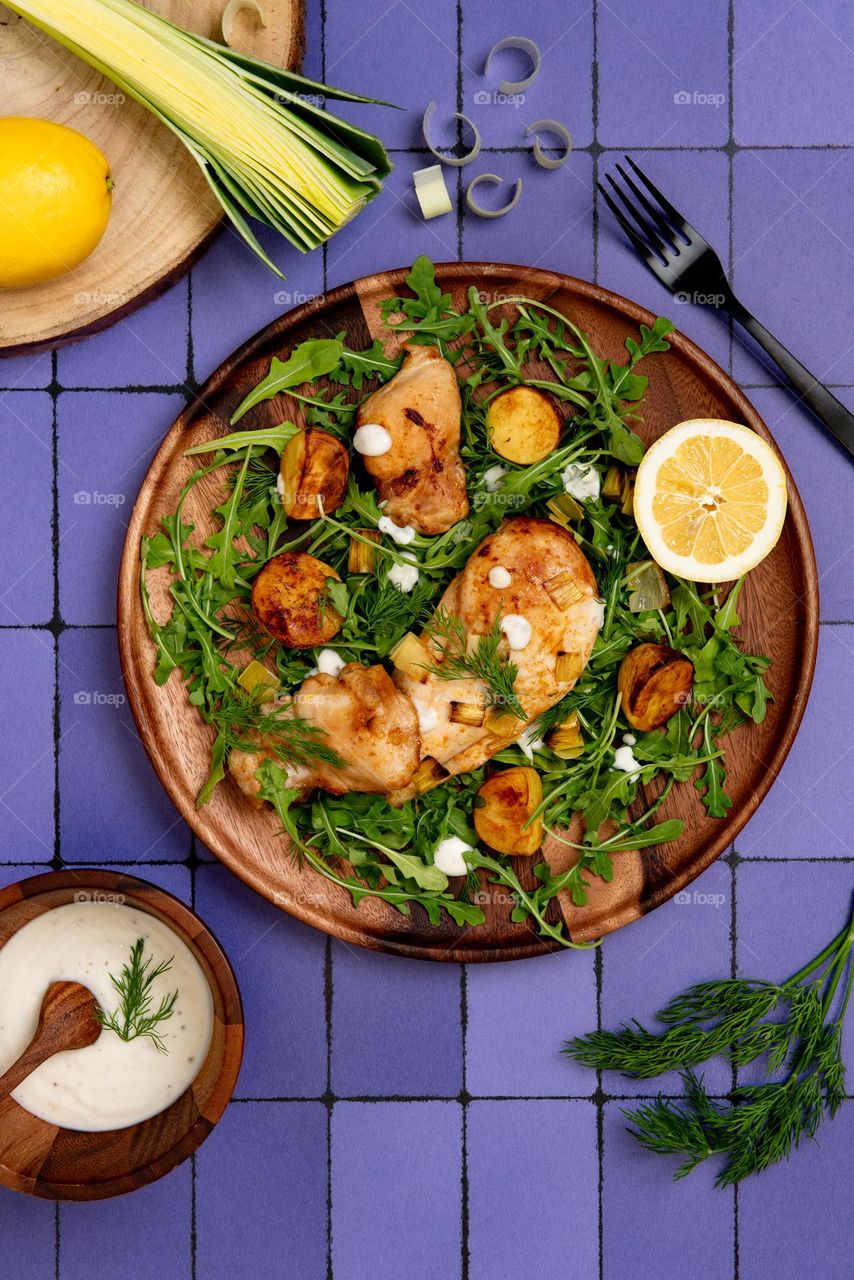 Flat lay of a fresh, healthy meal consisting of leeks, baby arugula, roasted chicken and potatoes with a lemon dill yogurt sauce on a purple tile surface 