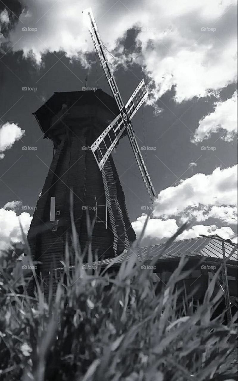 Black and white photo.  Bottom view.  In the foreground, grass through which you can see: an old wooden mill and a sky with clouds