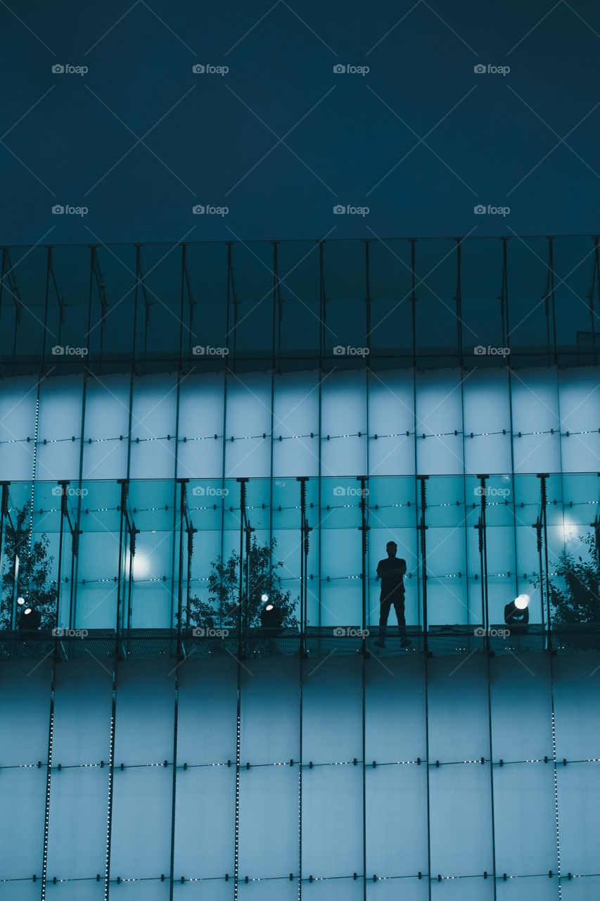 Silhouette of man standing on backlit glass construction in a city center at night