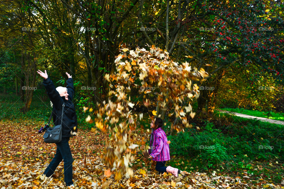 Grandma is throwing a pile of leafs up in the air, the little girl is walking under it