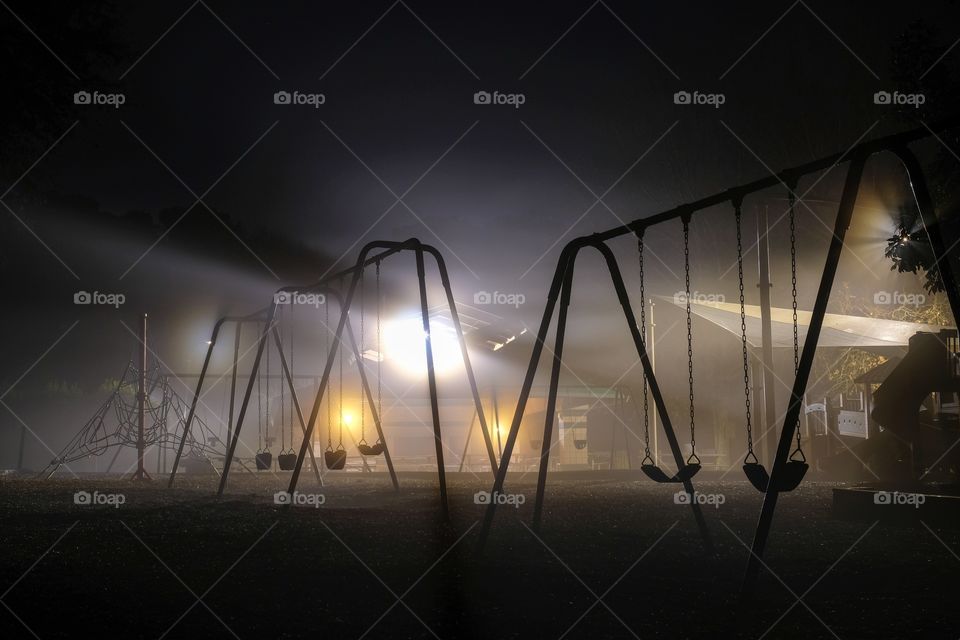 Swing sets backlit by streetlights on a dark foggy morning. Lake Benson Park in Garner North Carolina. 