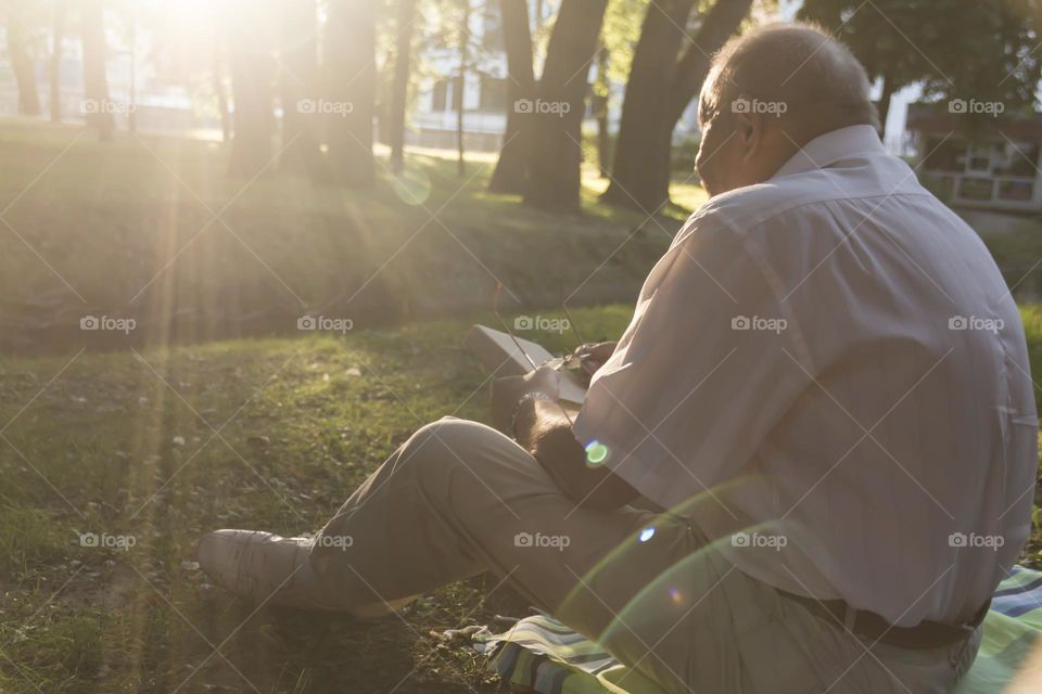 An elderly man of retirement age with wrinkles sits on the grass in the park and reads a book.