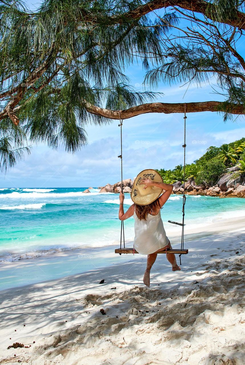 Rear view of young woman on wooden swing on an idyllic tropical sandy beach with turquoise ocean