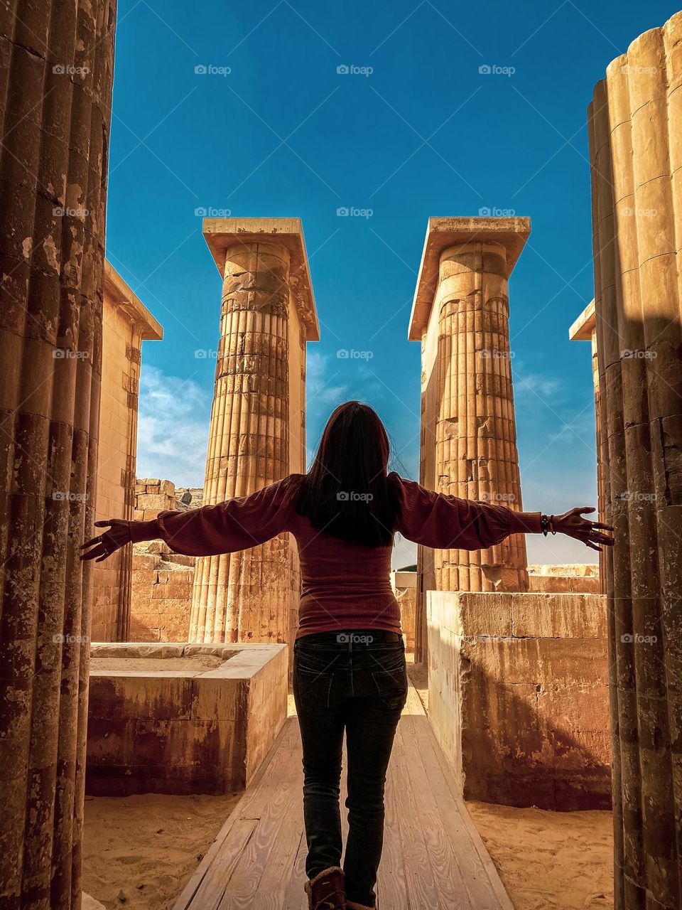 A woman walking in a pathway between columns at the Saqqara Archeological Complex. A sunny day with bright skies. Shadows add more cinematic look to the shot.