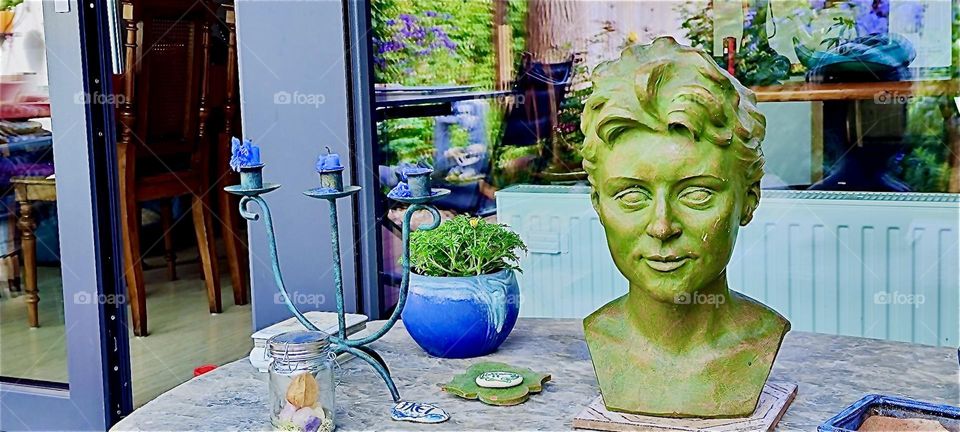 This beautiful bronze bust has been brought out into the garden today in rural “Bavaria”, Germany where it sits on a table with a candelabra and a potted plant. 2024. Hypnotic Productions