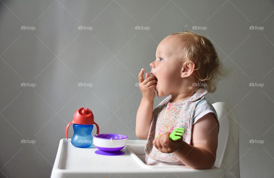 Little baby girl sitting in a feeding chair and eating porridge 