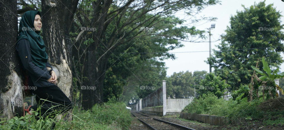 seorang wanita yang sedang bersandar pada pohon tua dengan pemandangan rel kereta api