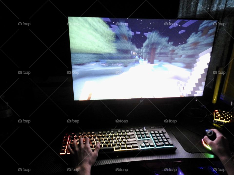 Teenager boy playing Minecraft on the gaming computer seen from his left side and close-up of his hand playing at his computer desk in a dark room.