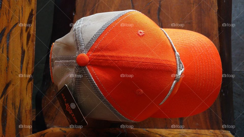 Sobre um banco de madeira, um boné na cor laranja, com telinha e bordados em cor prata envelhecida.