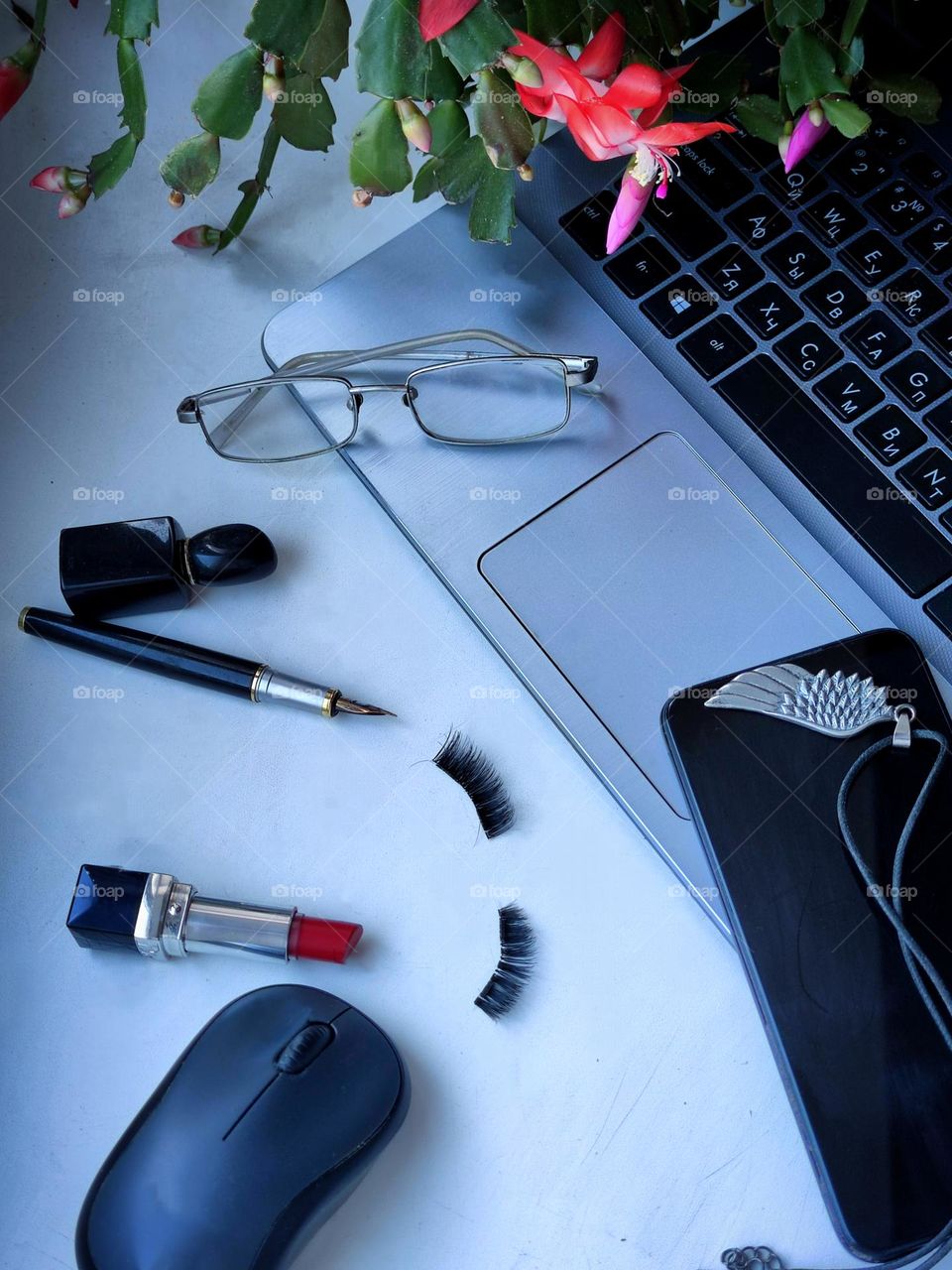 On a white surface is a part of a laptop on which leaves and flowers of a cactus fall. On the corner of the laptop are glasses and a phone with an pendant. Nearby on the surface are: a bottle of perfume, a pen, red lipstick, artificial eyelashes