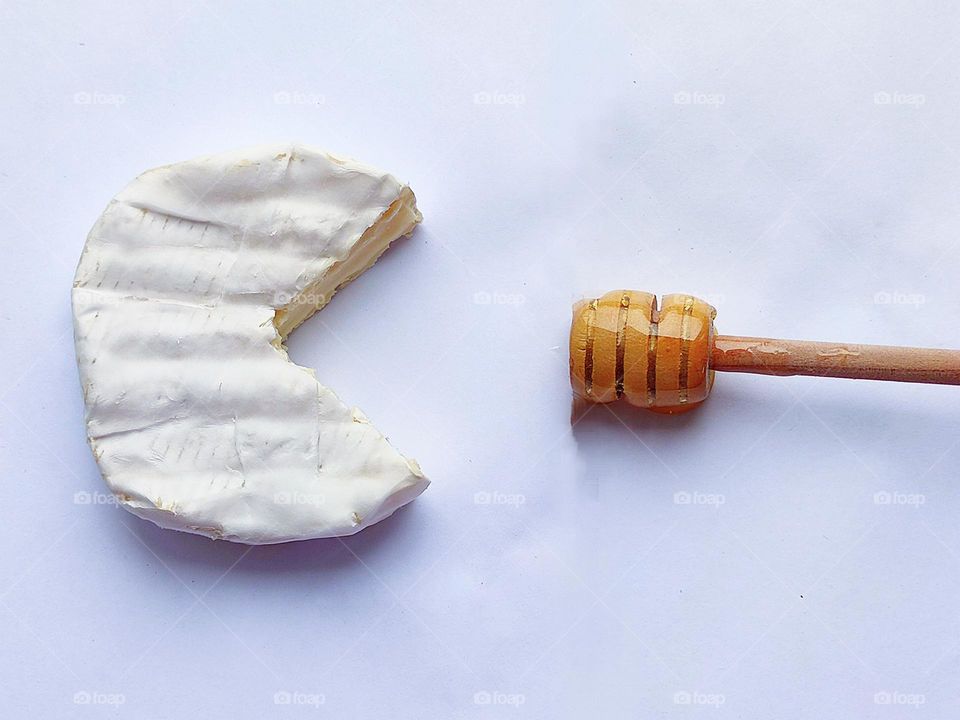 White colour.  White Brie cheese lies on a white background, part of which is cut off.  Not far from the cheese lies a wooden spoon with honey