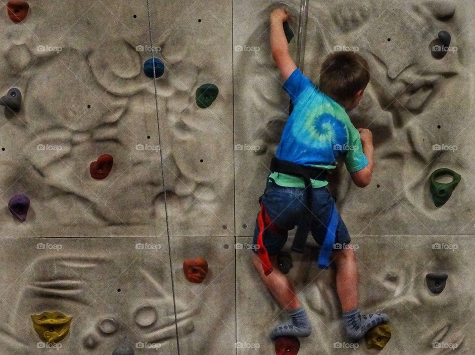 Young Boy Climbing A Rock Wall. Boy In Harness Scaling A Wall

