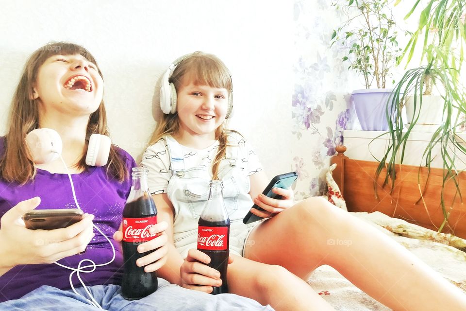 two teenage girls with a smartphone and headphones. they listen to music, drink Coca-Cola and laugh.