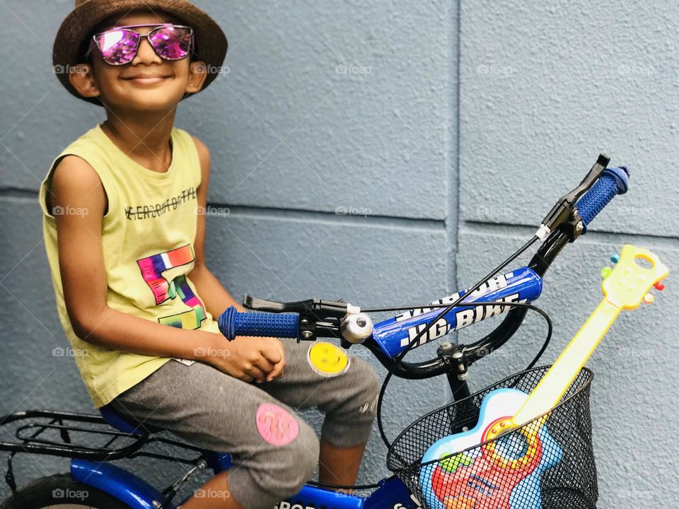 A funky and crazy style boy sitting on bicycle with guitar and eyeglasses and hat on head