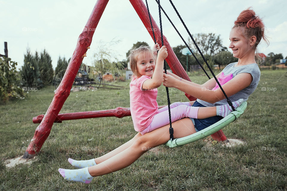 Teenage girl playing with her younger sister in a home playground in a backyard. Happy smiling sisters having fun on a swing together on summer day. Real people, authentic situations