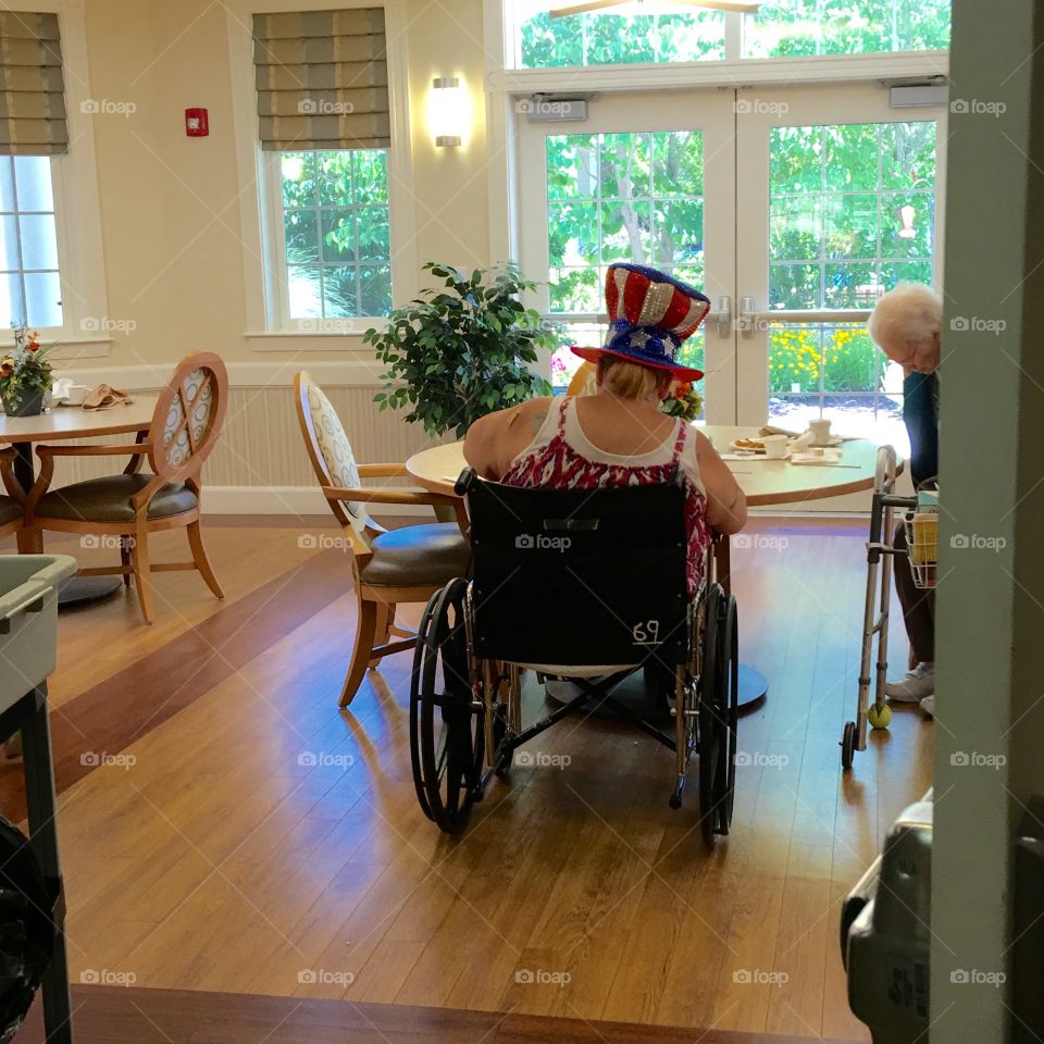 Nursing home wheelchair patient sitting alone in dining area, wearing patriotic hat.