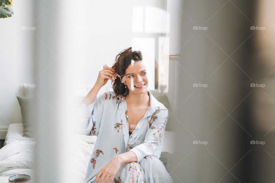 Young brunette woman in blue pajamas doing face massage with stone roller sitting on bed at home