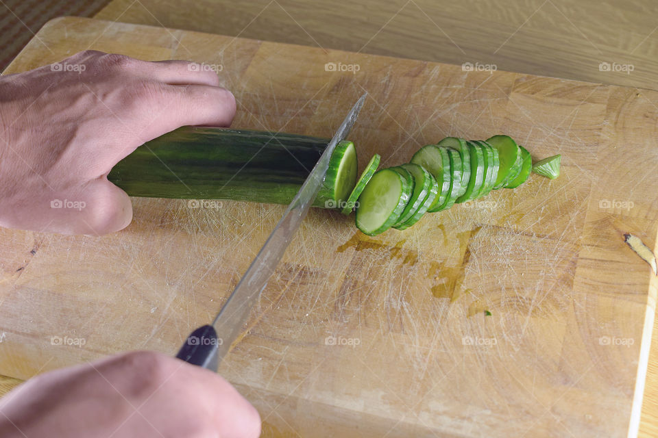Man cuts fresh cucumber to slices on a wooden cutting board.