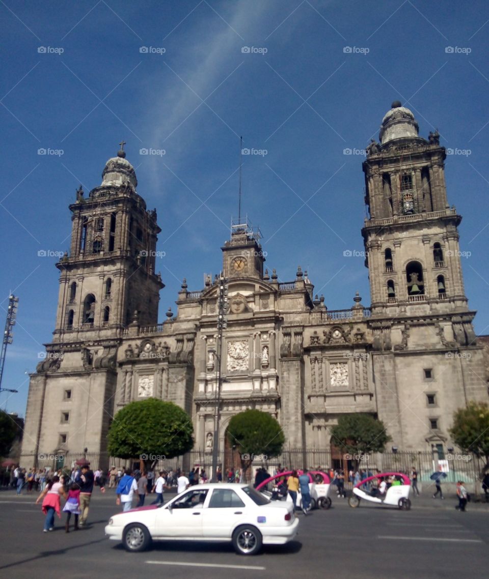 Edificio catedral de la cuidad de México. frente al zócalo capitalino. con autos pasando por enfrente, gente cruzando. de fondo el cielo azul.